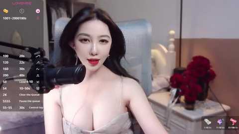 Media: A video of a pale-skinned woman with long black hair, wearing a low-cut, light-colored top, sitting in front of a camera. The background features a bedroom with a bed, nightstand, and red flowers.