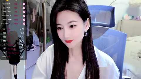 Media: Video of a fair-skinned Asian woman with long black hair, wearing a white blouse and red lipstick, seated in a room with a blue chair and cluttered background.