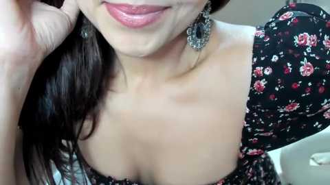 Media: A close-up video of a woman with light skin and long dark hair, wearing a black floral dress with a plunging neckline, large dangling earrings, and a soft smile.