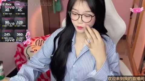 Media: Video of an East Asian woman with long black hair, wearing glasses, a striped shirt, and a ring, seated in a pink chair. Background includes a pink wall, a stuffed animal, and a digital screen displaying chat messages.