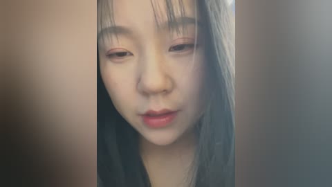 Media: Video of a young East Asian woman with pale skin, long black hair, and light pink eye shadow, looking contemplative, surrounded by blurred, beige walls.