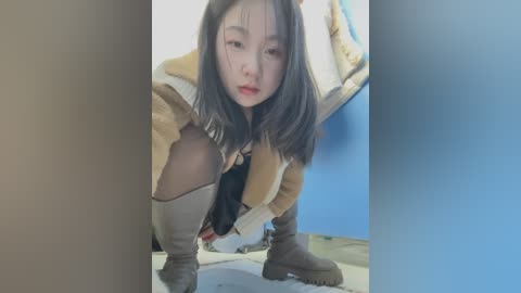 Media: Video of an Asian woman with long black hair, wearing a beige jacket, black top, and grey knee-high boots, squatting with her knees wide apart in a dimly lit room with blue walls.