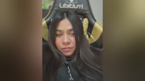 Video of a young Asian woman with long black hair, wearing a black hoodie, sitting on a black and gold UFC gaming chair. Her expression is calm and focused.