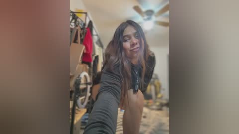 Video of a smiling woman with long, dark hair, wearing a black top, leaning forward, taking a selfie in a cluttered bedroom with clothes hanging on racks, a fan on the ceiling, and a bicycle in the background.