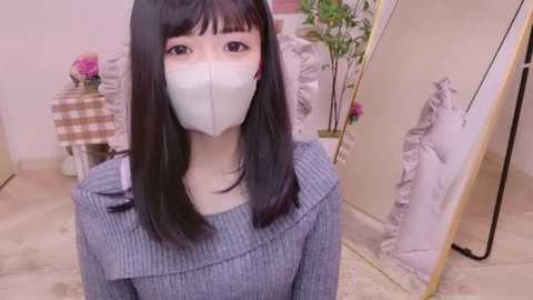 Media: Video of a young East Asian woman with straight black hair, wearing a gray ribbed sweater, white face mask, and a pink hair clip, standing in a cozy, minimalistic bedroom with a mirror, floral decor, and white bedding.