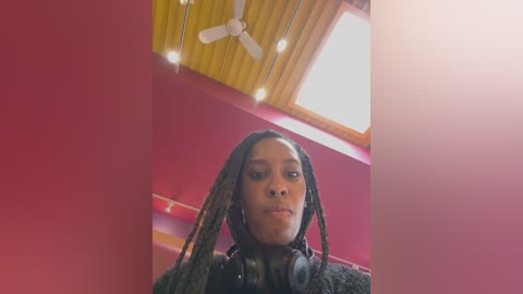 Media: Video of a young Black woman with braided hair, wearing headphones, standing in a room with red walls, wooden ceiling, and a skylight.