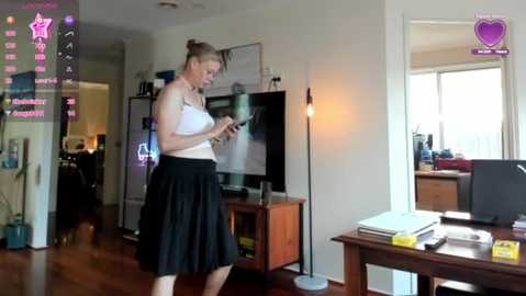 Media: A video of a slim, blonde woman in a white tank top and black skirt, typing on a tablet, in a modern living room with a flat-screen TV, wooden desk, and window.