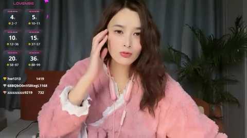 Media: Video of an Asian woman with long brown hair, wearing a pink fuzzy robe, sitting indoors with a potted plant in the background. She holds a phone to her ear. Live stream overlay shows streaming stats.