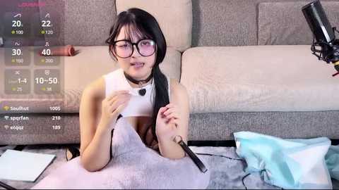 Media: Video of an Asian woman with glasses, light skin, small breasts, and black hair, lying on a gray couch, holding a microphone, surrounded by clothes and a laptop.