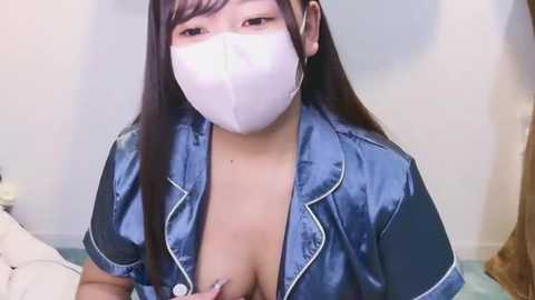 Media: Video of a young Asian woman with long black hair, wearing a blue satin robe with white trim, and a white face mask. She has a fair complexion and medium-sized breasts, partially visible. Background shows a simple indoor setting with a white wall and a bed.