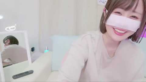 Media: Video of a smiling Asian woman with a straight bob haircut, wearing a white blindfold and a beige top, sitting in a brightly lit, minimalist room with white walls and light-colored furniture.