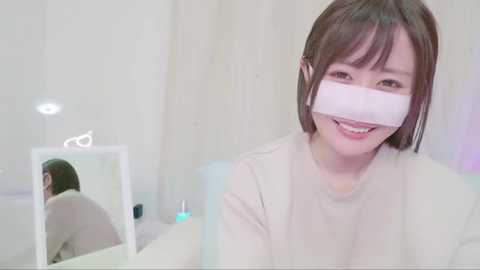 Media: Video of a smiling, light-skinned East Asian woman with straight brown hair, wearing a white face mask and a white long-sleeved shirt, standing in a minimalistic, white-walled room with a vanity mirror reflecting her back.