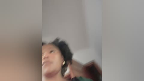 Media: Blurry video of a woman with dark skin, curly hair, and earrings, caught mid-movement, in a dimly lit room with white walls.