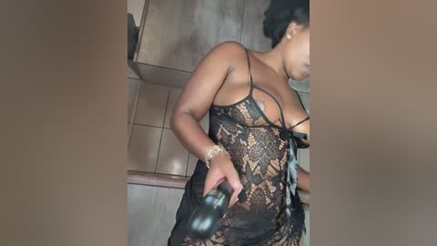 Media: Video of a dark-skinned woman with large breasts, wearing a sheer black lace lingerie top, standing in a kitchen with tiled walls.