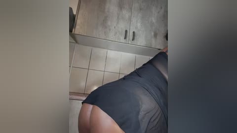 Media: Video of a woman in a sheer black bodysuit, bending over in a kitchen with tiled backsplash and wooden countertops, focusing on her large, round buttocks.