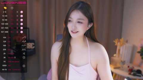 Media: A young Asian woman with long brown hair, wearing a pink tank top, smiles while looking at a camera in a softly lit room.