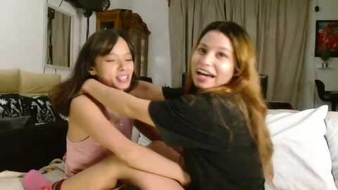 Media: Video of two Asian women hugging in a cozy living room; one wears a pink tank top, the other a black shirt; beige curtains, wooden furniture, and a black sofa in the background.