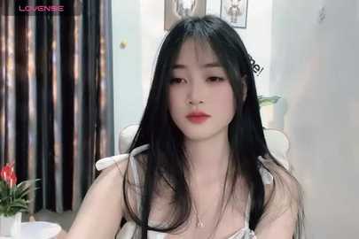 Media: Video of a young Asian woman with long black hair, fair skin, and full lips, wearing a white top, sitting indoors with a floral arrangement in the background.