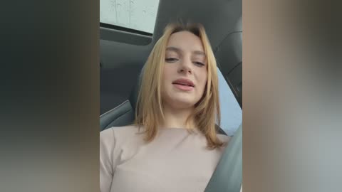 Media: A video of a young Caucasian woman with straight blonde hair, wearing a light pink top, reclining in a car's grey leather seat.