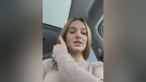 Media: Video of a young woman with light skin and shoulder-length blonde hair, wearing a beige long-sleeved top, sitting in a car with a grey interior, smiling and adjusting her hair.