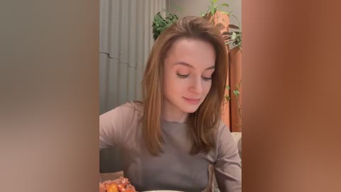 Media: Video of a fair-skinned, red-haired woman with shoulder-length hair, wearing a gray top, eating popcorn in a cozy, dimly lit room with greenery and a wooden shelf in the background.