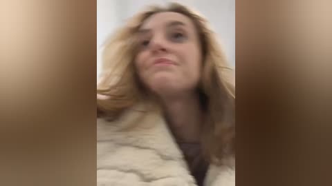 Media: Video of a blonde woman with a fair complexion and a fur coat, her hair blowing in the wind. The background is blurry, emphasizing her face and hair.