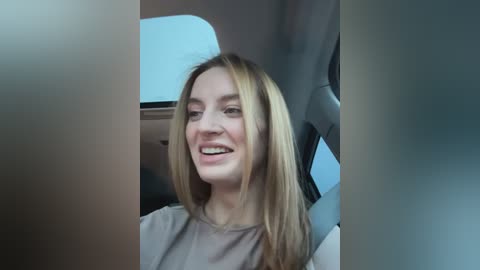 Media: Video of a smiling, fair-skinned, blonde woman with shoulder-length hair, wearing a light grey top, seated in a car with grey upholstery.