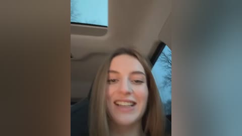 Media: Video of a smiling young woman with long blonde hair, sitting in a car with a window view of a leafless tree outside.