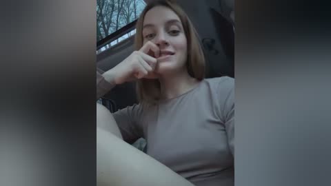 Media: Video of a Caucasian woman with light skin and shoulder-length brown hair, wearing a light grey long-sleeved shirt, sitting in a car, looking relaxed, with bare tree branches visible outside.