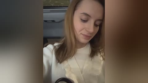 Media: A video shows a young woman with fair skin, light brown hair, and a white sweater, sitting in a car. She has a slight smile and is looking down. The background features a blurred view of the car's interior.