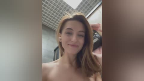 Media: Video of a topless woman with light skin and shoulder-length, light brown hair, smiling, in a bathroom with a grid ceiling and tiled walls, holding a black hairbrush.