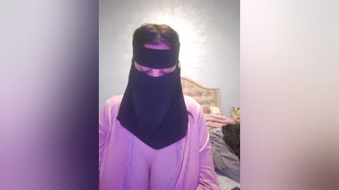 Media: Video of a person wearing a black mask with a white blindfold, standing in a bedroom with a headboard and bed visible in the background.