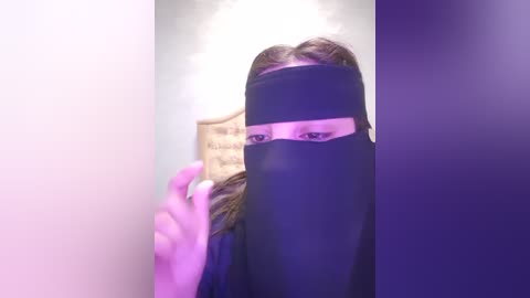 Media: A video of a woman with light skin and shoulder-length brown hair, wearing a black hijab and holding a wooden frame. She is in a room with a soft pink and purple light gradient.