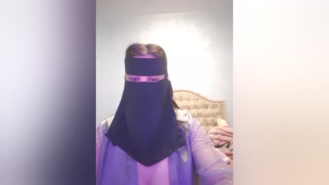 Media: A video of a person wearing a black niqab with a pink headband, standing in a dimly lit room with a white wall and a beige wicker headboard.