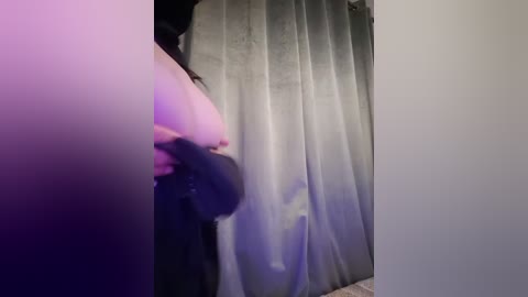 Media: Video of a nude woman with large breasts and fair skin, partially obscured by a grey curtain in a dimly lit room, with purple lighting.