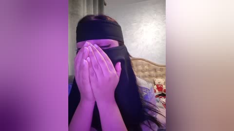 Media: A video of a person with long, dark hair wearing a black blindfold, holding their hands to their face in a bedroom with a bed, teddy bear, and textured wall.