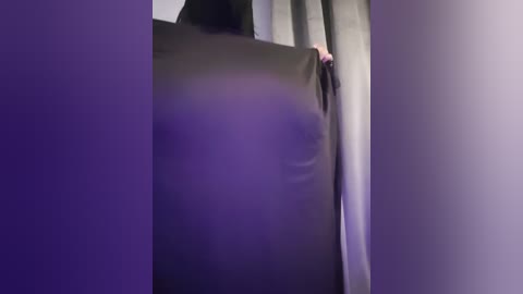 Media: Video of a woman's hand holding a black curtain in a dimly lit room, with soft purple lighting creating a moody atmosphere.