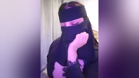 Media: Video of a woman with long, straight dark hair, wearing a black niqab and long-sleeved black dress, sitting with her head lowered, hand covering her face. The background is a plain, light-colored wall.