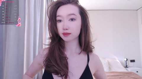 Media: Video of a young Asian woman with long, wavy brown hair, wearing a black spaghetti-strap top, standing in a bright, modern bedroom with white walls and minimalistic decor.