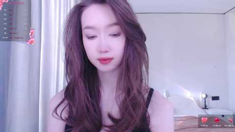 Media: Video of a young Asian woman with long, wavy brown hair, fair skin, and red lipstick, wearing a black top, in a modern bathroom with a white countertop and mirror.