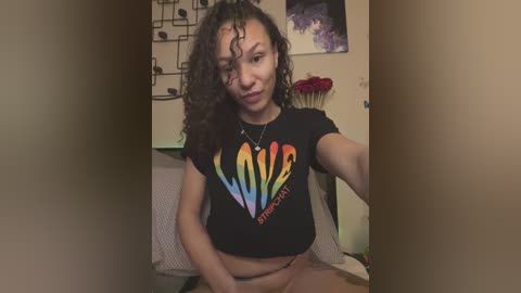 Media: Video of a young Black woman with curly hair, wearing a \"Love is Love\" t-shirt, smiling, sitting on a bed with a colorful wall and red flowers.