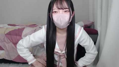 Media: Video of a young Asian woman with long black hair, wearing a white sailor school uniform, pink bow, and face mask, kneeling on a carpeted floor, in a bedroom with a patterned bedspread.