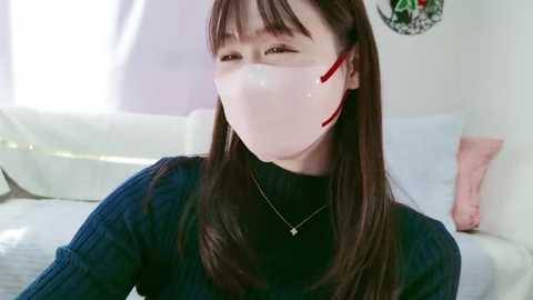 Media: Video of a young woman with straight brown hair, wearing a dark navy sweater and clear face mask with red ribbons. She's in a brightly lit room with white furniture and a decorative wall hanging.