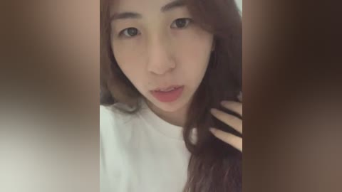 Media: Video of a young Asian woman with long, straight brown hair, light skin, and expressive eyes. She wears a white T-shirt, and her right hand is gently touching her hair. The background is blurred, suggesting an indoor setting.