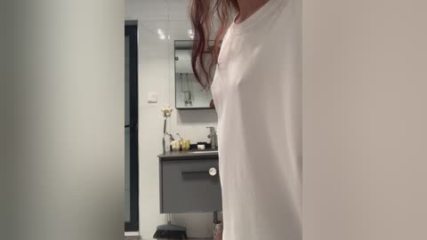 Media: Video of a slender, fair-skinned woman with auburn hair, wearing a loose white shirt, standing in a modern, minimalist bathroom with white tiles, a grey vanity, and a mirror.