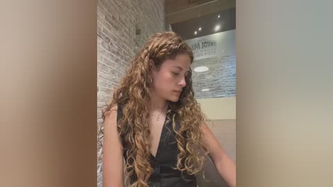 Media: Video of a young woman with long, curly, light brown hair in a black sleeveless dress, seated against a brick wall in a modern salon with a sign reading \"A Cut Above.\