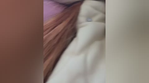 Media: A blurred video of a person's torso with light skin and long, reddish-brown hair, lying on a white bedsheet. The background is indistinct and muted.