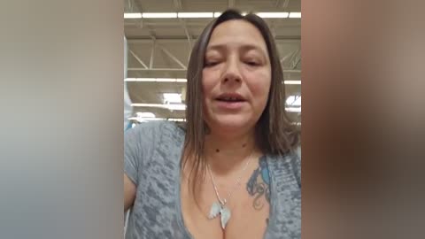 Media: Video of a middle-aged woman with shoulder-length brown hair, wearing a gray patterned top, and a necklace with a feather pendant, smiling in an indoor setting with a high ceiling and visible beams.