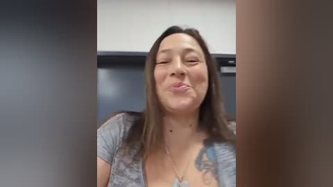 Media: Video of a smiling Asian woman with medium-length brown hair, wearing a gray T-shirt, making a kissy face indoors. Background features a blurred, dark room.