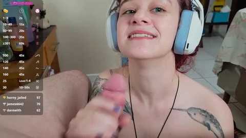 Video of a fair-skinned woman with short red hair, smiling, wearing large headphones, and holding a realistic, erect penis. Background shows a cluttered room with gaming equipment and a monitor displaying chat messages.